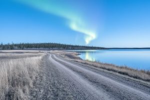 Wide prairie landscape in Manitoba at winter, with distant forest, a reflective lake, and faint aurora lights across the night sky.